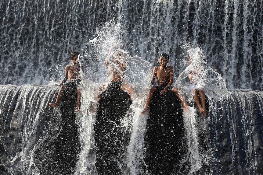 Boys sit under a dam with water spraying around.