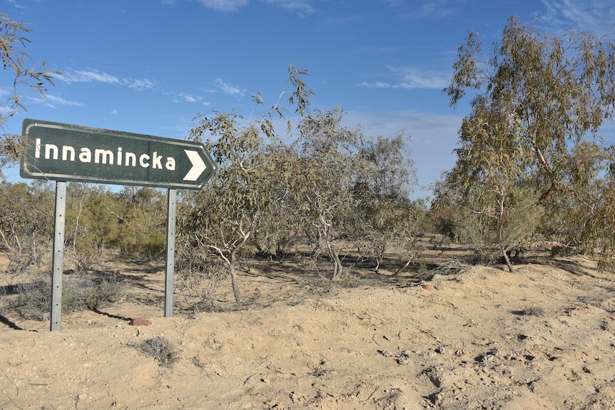 A green road sign surrounded by trees and sand.  The sign says Innamincka on it.