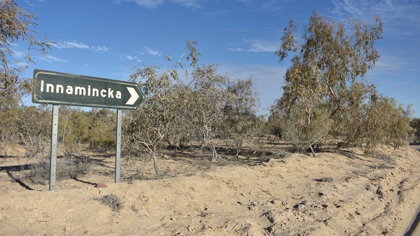 A green road sign surrounded by trees and sand.  The sign says Innamincka on it.