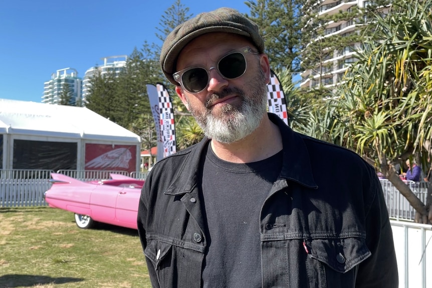 A man with a grey beard, round sunglasses and a hat standing in front of a pink car