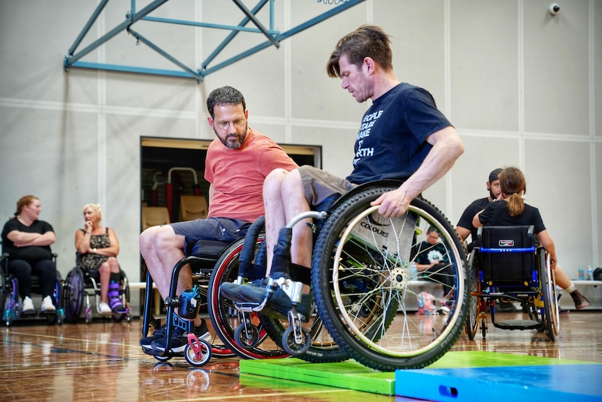 Two men sit in wheelchairs, with the man on the right showing the man on the left how to perform a manoeuvre.