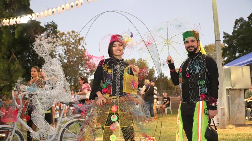 A man and a woman wear colourful recycled fashion next to a sculpture of a person on a bike a the Katherine Junk Festival. 