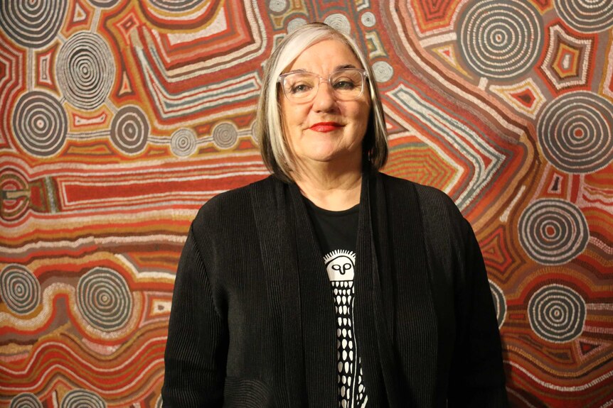 Margo, wearing lipstick, smiles in front of an Indigenous painting which is red and brown in colour.