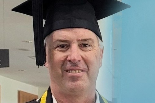 A man stands in university graduation gowns and cap.