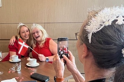 Two women wearing red tops and holding a red and white Danish flag smile as another woman takes a photo of them