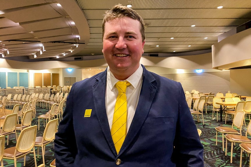 A man in a navy jacket with a yellow tie smiles, rows of empty cream chairs behind him.