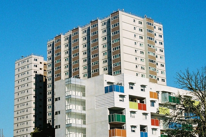 On a bright blue day, you view a brutalist public housing tower with a brightly-coloured contemporary tower in front of it.