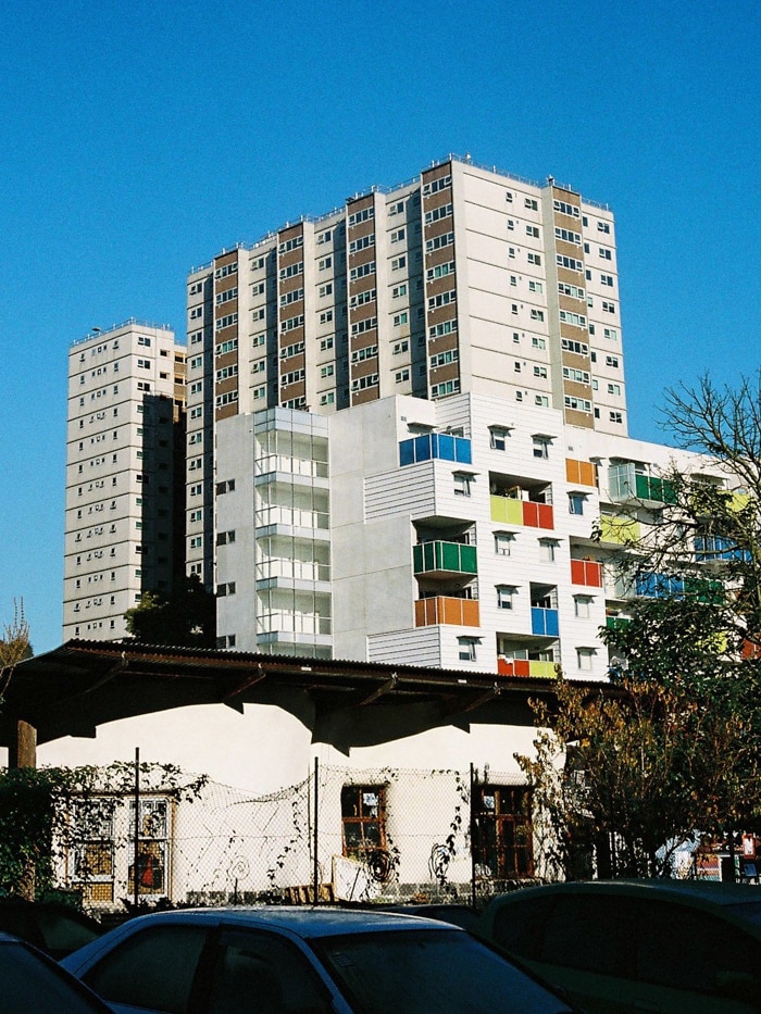 On a bright blue day, you view a brutalist public housing tower with a brightly-coloured contemporary tower in front of it.