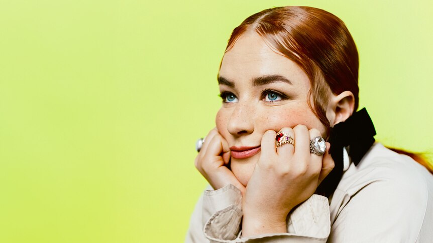 Mia Wray on a bright green backdrop sits with her head in her hands and her bright red hair slicked back.