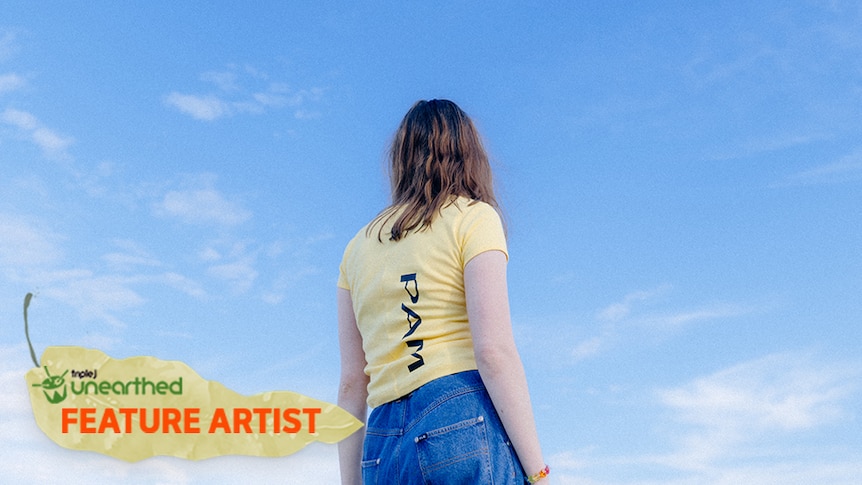 cookii, a woman with light brown hair and a yellow t-shirt, looks away from the camera in front of a bright blue sky.