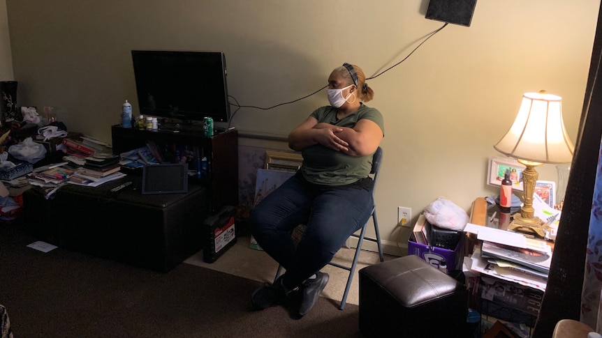 A woman sits on a folding chair in a cluttered, dark living room 