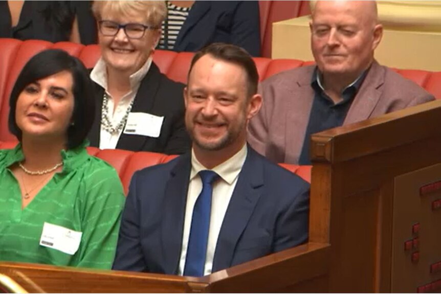 Ben Hood smiling sitting down in parliament with one person seated next to him two seated behind