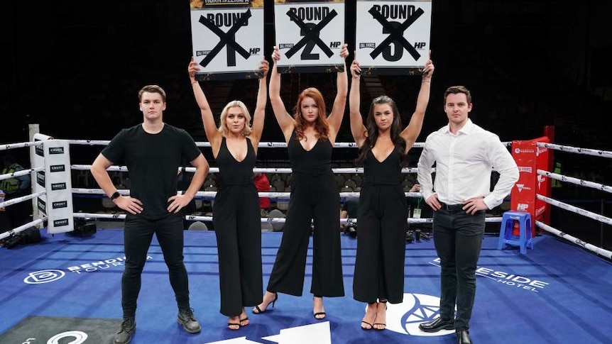 Two men and three women stand in a boxing ring holding round cards.