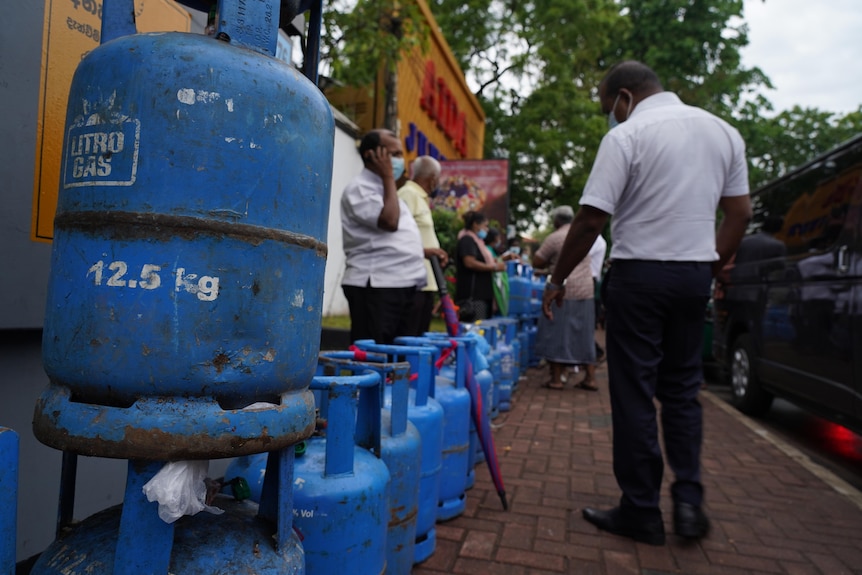 A close-up shows a blue gas bottle leaning on a stack of other gas bottles, with people lined up down the block