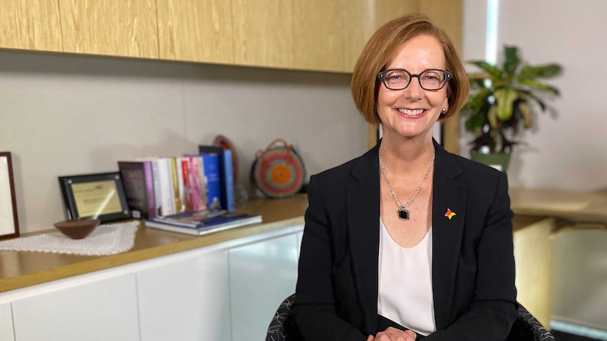 A woman with red hair and glasses sitting in an office with her legs crossed smiling.