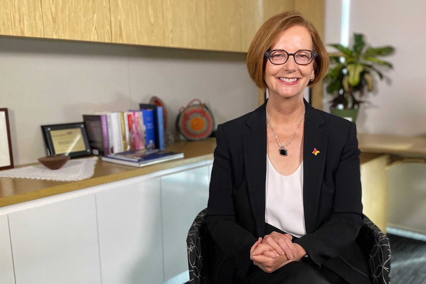 A woman with red hair and glasses sitting in an office with her legs crossed smiling.