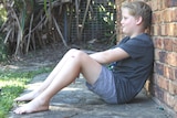 13yo Oliver Sun sits against the brick wall of his house gently smiling.