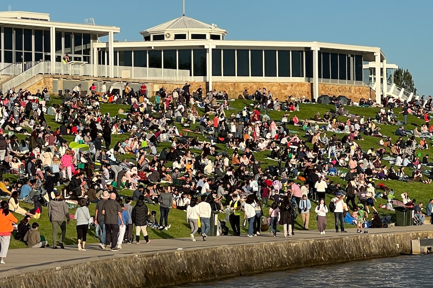 A wide image of people gathering on a hill behind a body of water. Some people walk on a path near the shore.