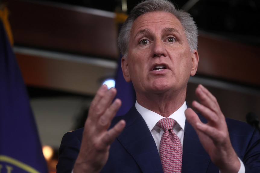 A man wearing a suit with a red tie holds gestures with his hands as he speaks.