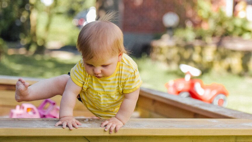 A toddler in a yellow play suit climbs carefully out of a sand pit