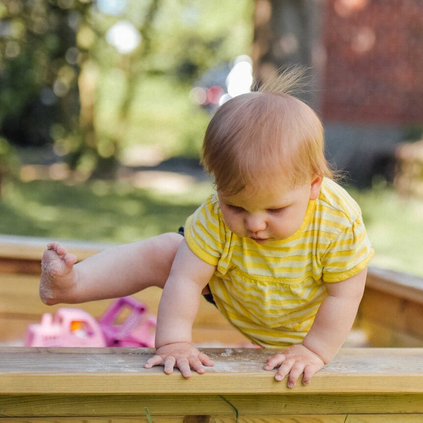 A toddler in a yellow play suit climbs carefully out of a sand pit