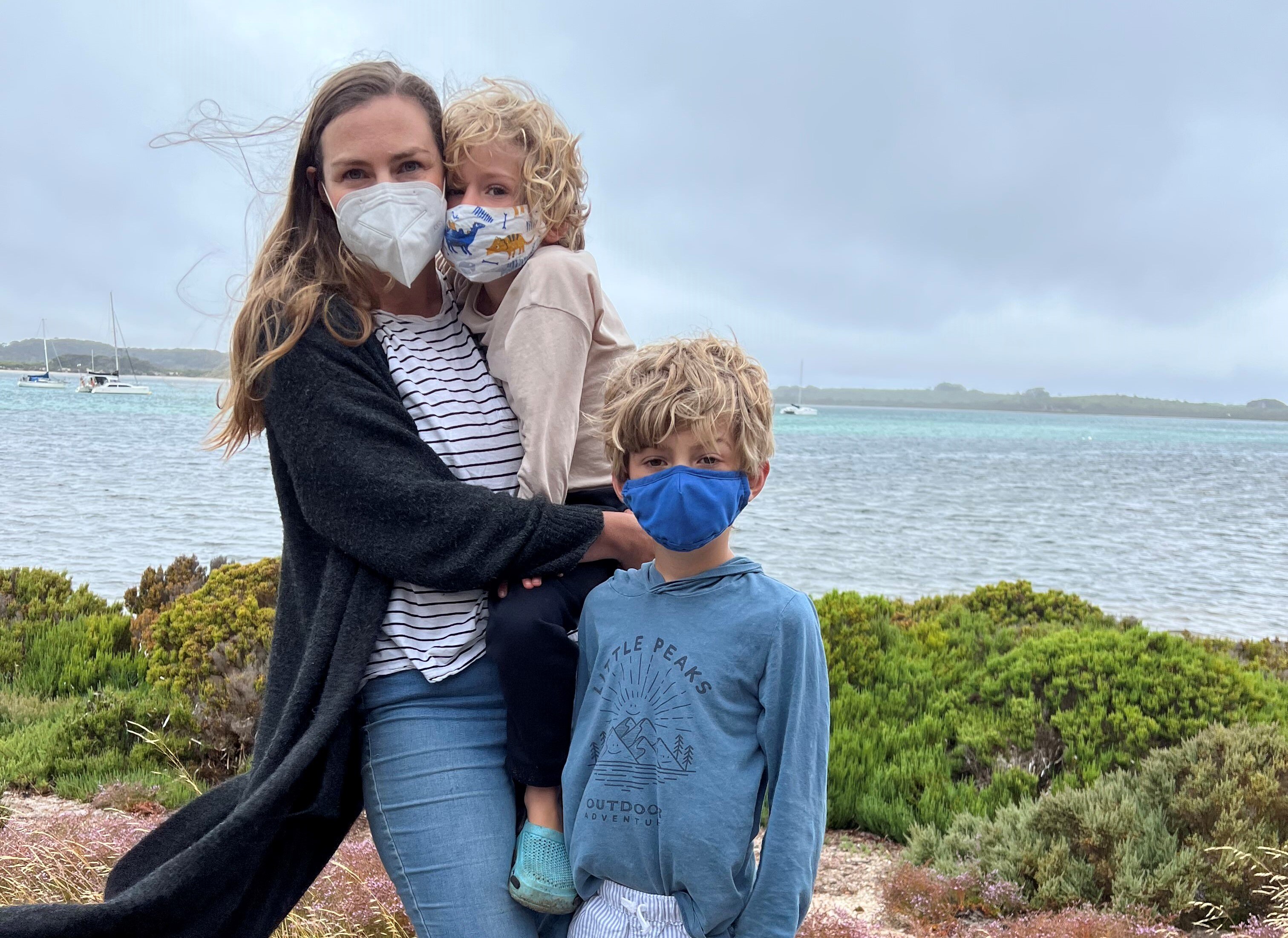 A woman holds a young boy and stands next to another in front of water. They're all wearing face masks. 