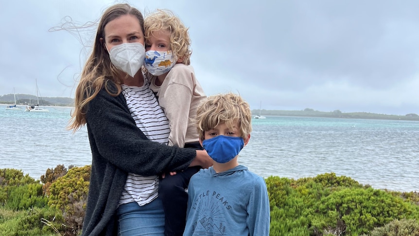 A woman holds a young boy and stands next to another in front of water. They're all wearing face masks. 