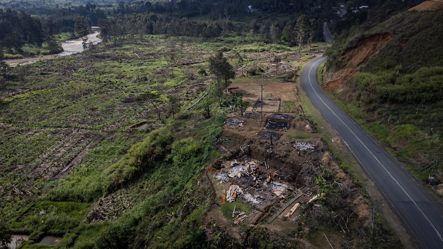 An aerial view shows the burnt remains of structures, next to a road heading down a hill towards a village