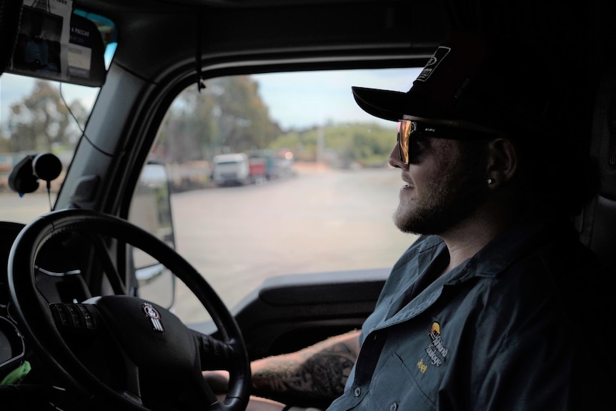 A man wearing sunglasses and a cap behind the steering wheel of a truck.