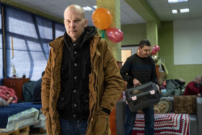 A man looks at the camera with a neutral expression, hands in his pockets. Behind him another man holds a cat carrier