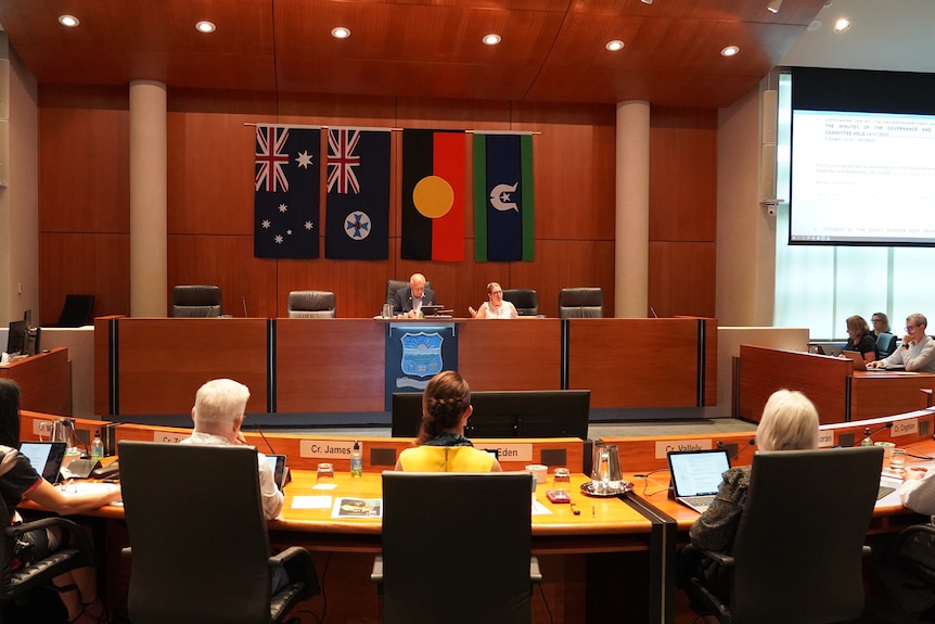 A council chambers, with many people sitting at their assigned desks.