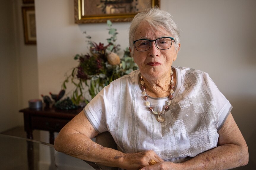 Helen wearing a white blouse and black-rimmed glasses sits with her arms in front in her home.