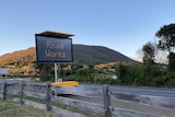 An electronic roadworks sign sits between a timber fence and a road with hills in the background.