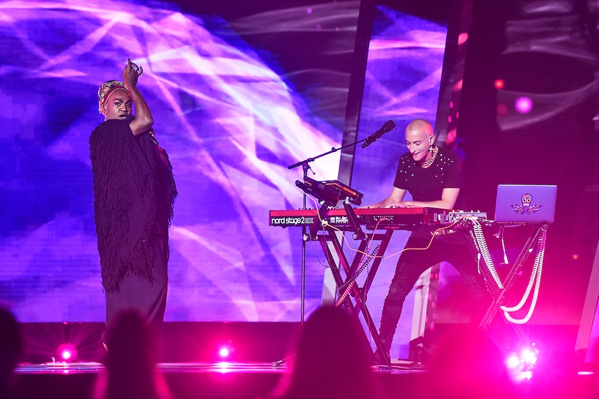 A man poses and sings next to another man playing the keyboard, both are performing on purple hued stage in front of screens.