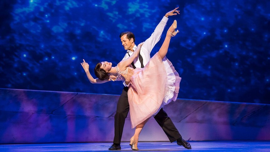 A dancer in a pink dress is dipped by a dancer in a suit, set against a deep blue background