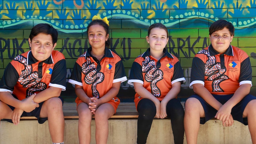 Two boys and two girls wearing indigenous themed school uniforms sitting on a bench in front on an indigenous mural.