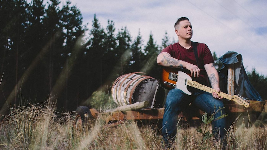 Gareth Leach holding a guitar and sitting on a rusted trailer.