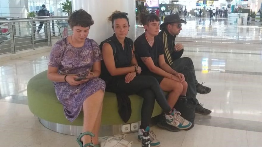 Four people, three women and a man, sit in an airport. One is looking at a phone.