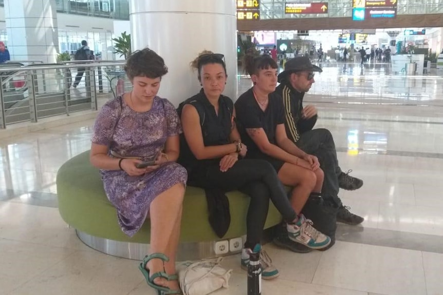 Four people, three women and a man, sit in an airport. One is looking at a phone.