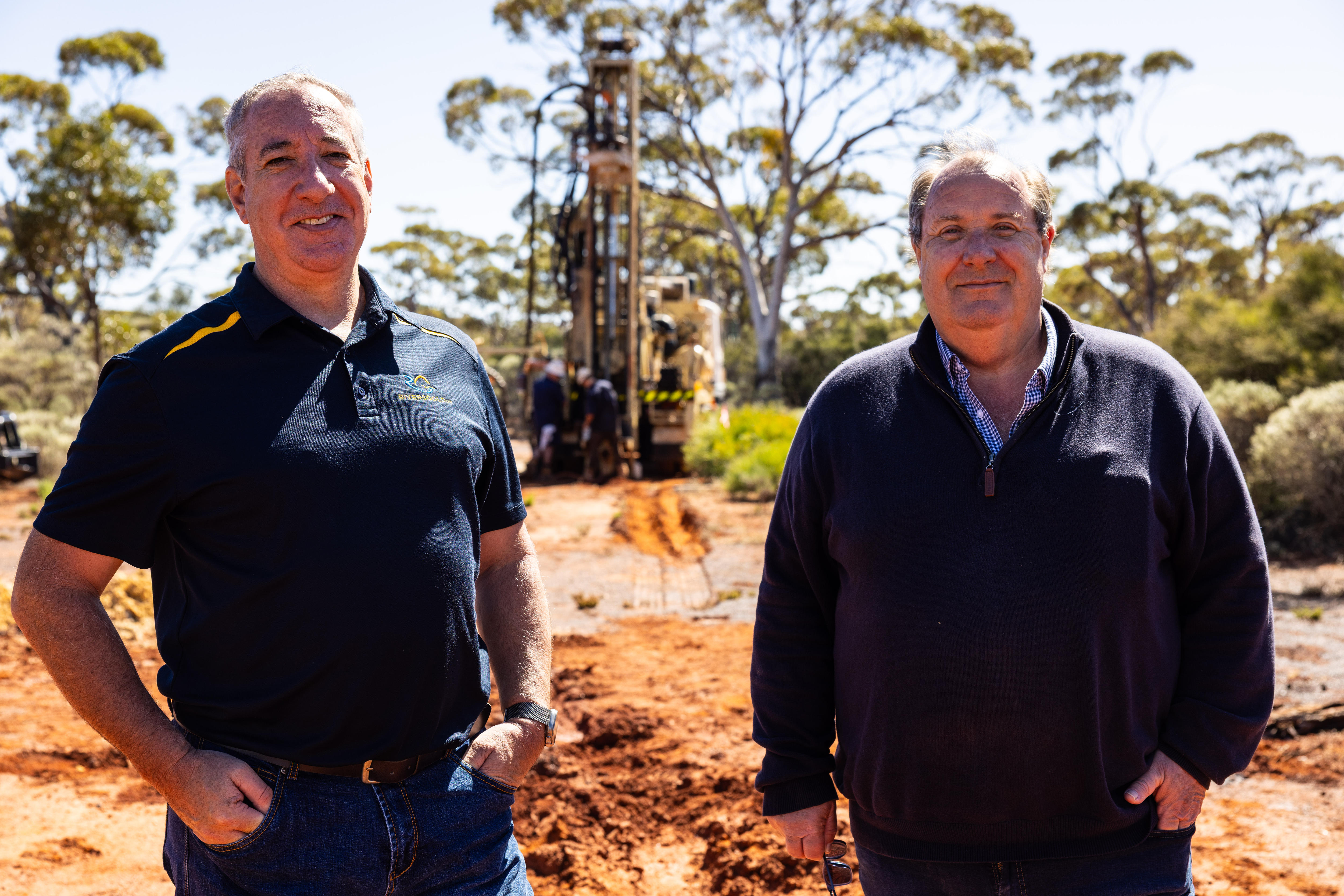 Two men standing in front of a drilling rig in bushland.  