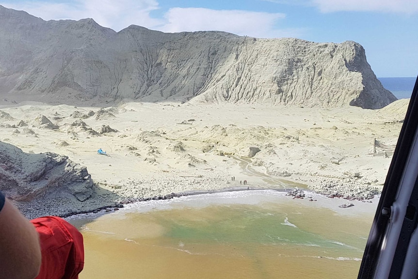 A view of White Island from the door of a chopper. The crater is grey with volcanic ash with a number of people on the beach.