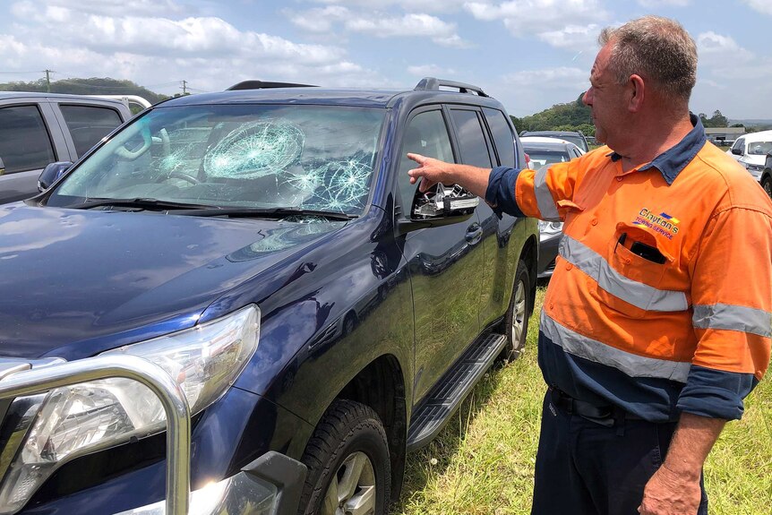 A man points at a four-wheel-drive with a hail damaged windscreen and dents.