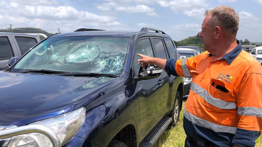 A man points at a four-wheel-drive with a hail damaged windscreen and dents.