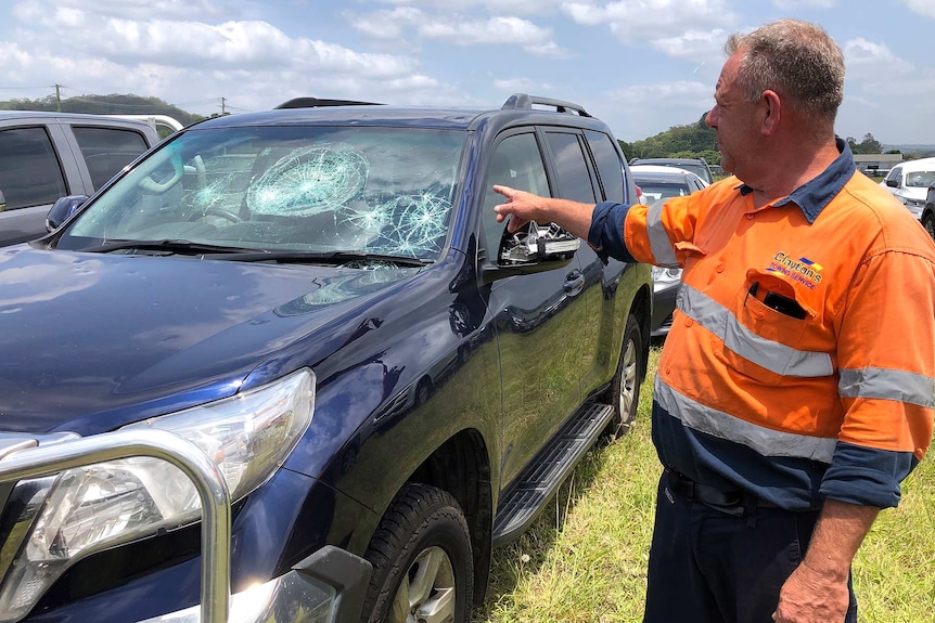 A man points at a four-wheel-drive with a hail damaged windscreen and dents.