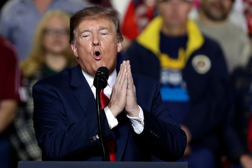 A man holds his hands together as he speaks to a large crowd of supporters at a rally.