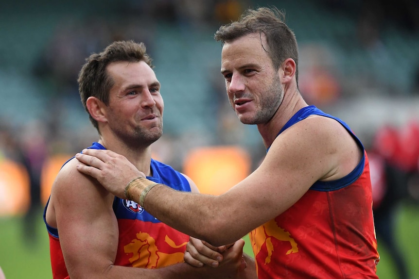 Two male AFL players smile and shake hands as they celebrate a win.