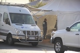 A police van sits parked on the side of a road next to a white tent with a forensics officer standing inside.