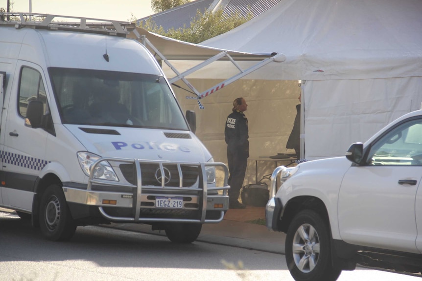 A police van sits parked on the side of a road next to a white tent with a forensics officer standing inside.