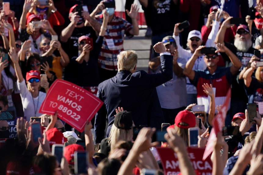 A man with blonde hair in a suit stands in a crowd of people with his fist in the air