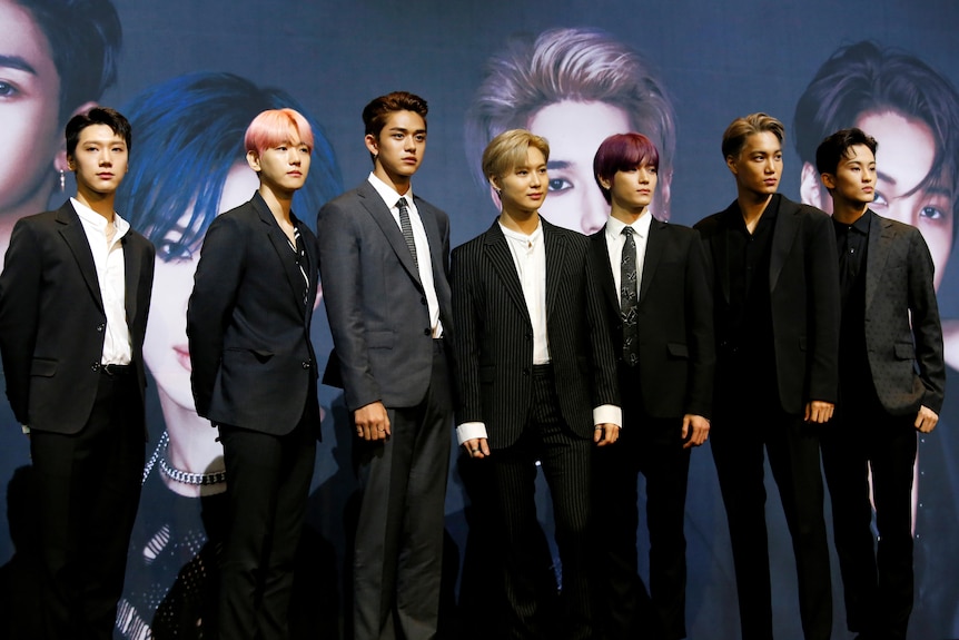 Seven men wearing suits stand in front of a giant poster of their own faces
